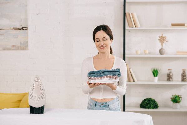 happy housewife holding ironed clothes while standing near ironing board