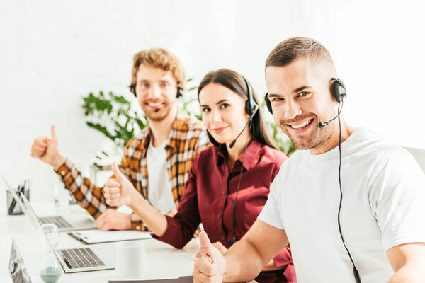 избирательный фокус счастливого брокера, показывающего большой палец рядом с коллегами в офисе
 