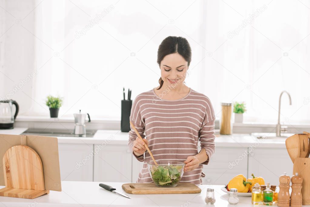 young, attractive woman mixing fresh vegetable salad while standing at kitchen table