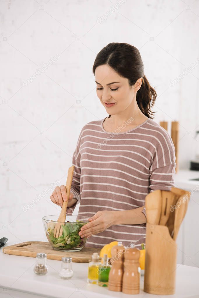 beautiful young woman mixing fresh vegetable salad while standing at kitchen table