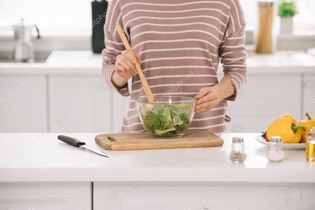 partial view of woman mixing fresh vegetable salad while standing at kitchen table