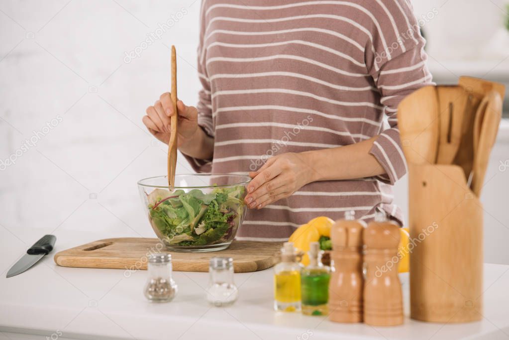 cropped view of woman mixing fresh vegetable salad while standing at kitchen table