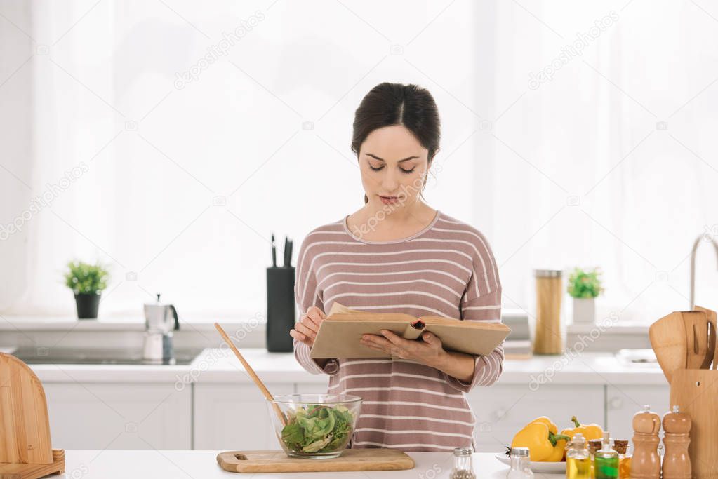 attractive, young woman reading receipt book while standing at kitchen table near bowl with vegetable salad