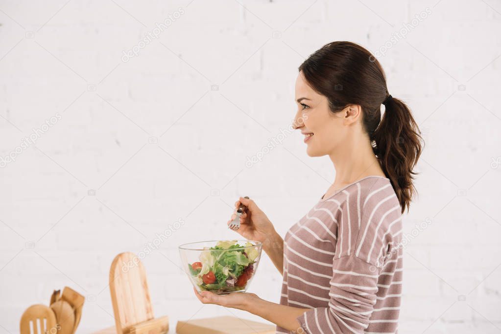 side view of attractive, smiling woman holding bowl with fresh vegetable salad