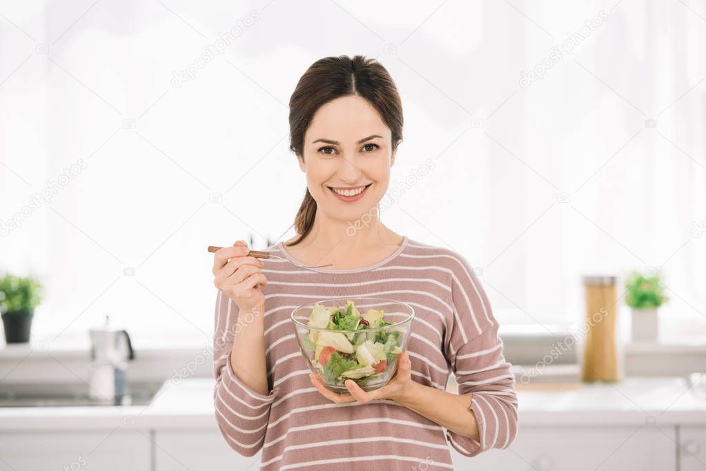 attractive, smiling woman looking at camera while holding bowl with vegetable salad