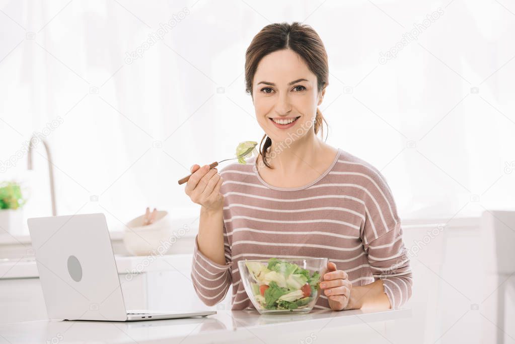 young, happy woman looking at camera while sitting at table near laptop and eating vegetable salad