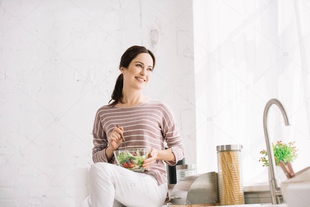 happy young woman looking away while sitting on kitchen table and holding bowl with vegetable salad