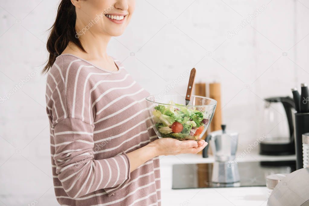 cropped view of young, smiling woman holding bowl with vegetable salad