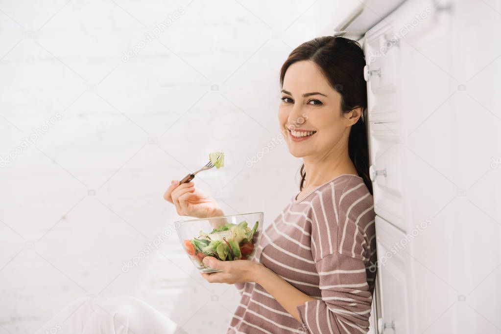 happy young woman looking at camera while sitting on floor in kitchen and eating vegetable salad