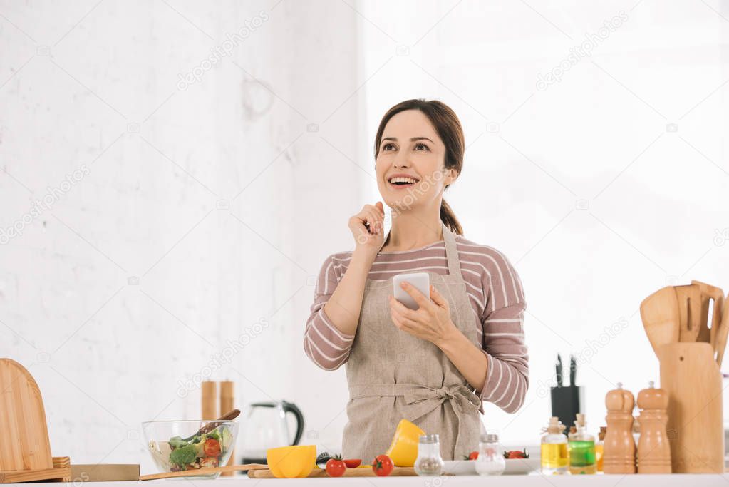 happy young woman in apron holding smartphone while standing at kitchen table near fresh vegetables