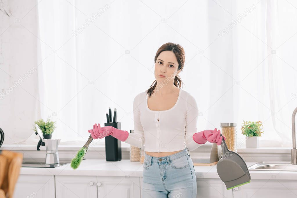 pensive housewife holding scoop and brush while looking at camera in kitchen
