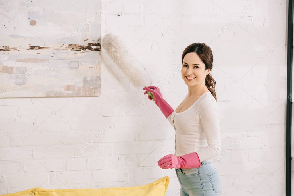 cheerful housewife smiling at camera while cleaning picture on wall with dusting brush