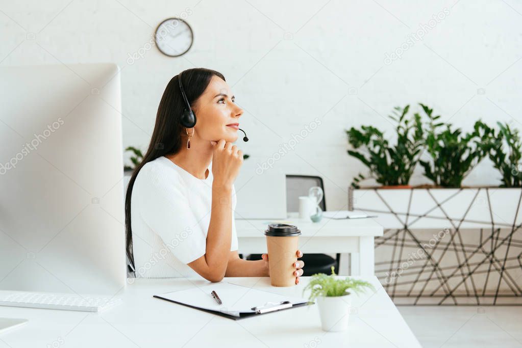 smiling broker in headset sitting and holding paper cup in office 