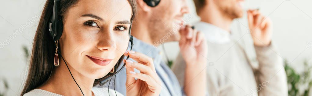 panoramic shot of happy broker touching headset near coworkers in office 