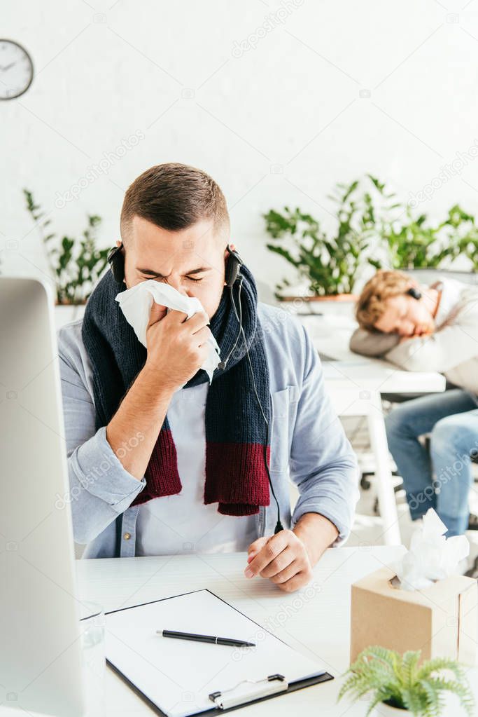 selective focus of sick broker sneezing in tissue near coworker in office 