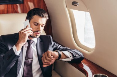 Takım elbiseli yakışıklı iş adamı akıllı telefondan konuşuyor ve özel uçakta kol saatine bakıyor. 