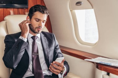 Takım elbiseli yakışıklı iş adamı müzik dinliyor ve özel uçakta akıllı telefon kullanıyor. 