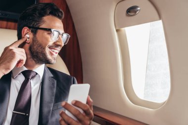 Gülümseyen iş adamı müzik dinliyor ve özel uçakta akıllı telefon kullanıyor. 