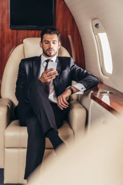 Özel uçakta akıllı telefon kullanan takım elbiseli yakışıklı iş adamının seçici odağı. 