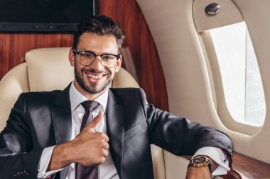 handsome businessman in suit smiling and showing thumb up in private plane 