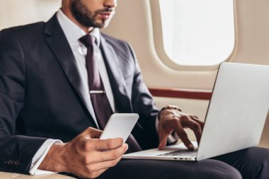 cropped view of businessman in suit using laptop and smartphone in private plane  clipart
