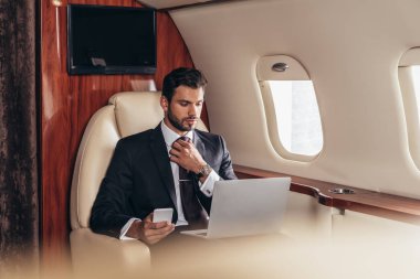 selective focus of businessman in suit using laptop and holding smartphone in private plane  clipart