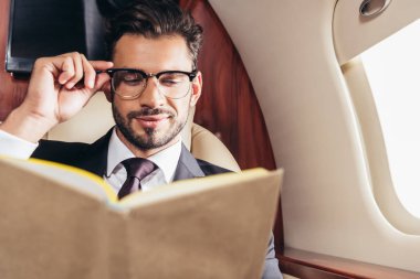 Takım elbiseli yakışıklı iş adamının seçici odak noktası özel uçakta kitap okuması. 