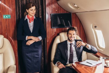 flight attendant in uniform looking at smiling businessman in private plane  clipart