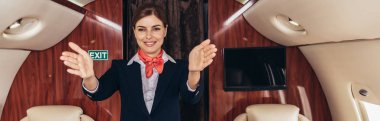 panoramic shot of smiling flight attendant in uniform with outstretched hands in private plane  clipart