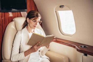 Takım elbiseli çekici bir iş kadını özel uçakta kitap okuyor. 
