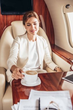 Takım elbiseli gülümseyen iş kadını özel uçakta kahve içiyor. 