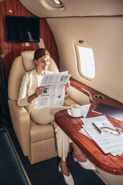 Takım elbiseli gülümseyen iş kadını özel uçakta gazete okuyor. 