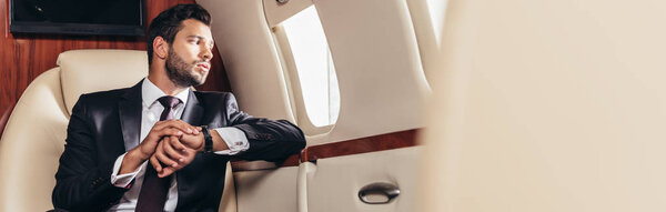 panoramic shot of handsome businessman in suit looking through window in private plane 