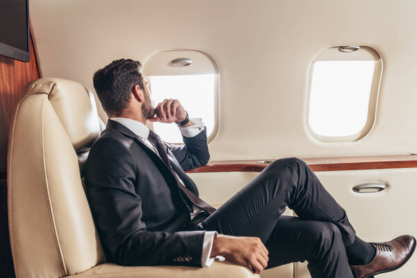 задний вид бизнесмена в костюме, смотрящего в окно в частном самолете
 