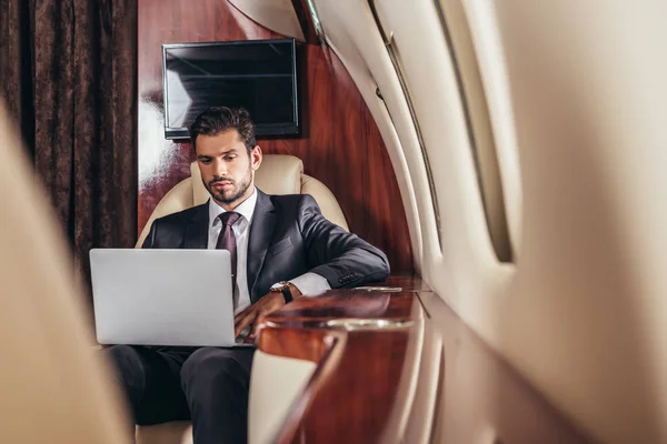 在私人飞机上使用笔记本电脑的英俊潇洒的穿着西装的商人的首选焦点 — 图库照片