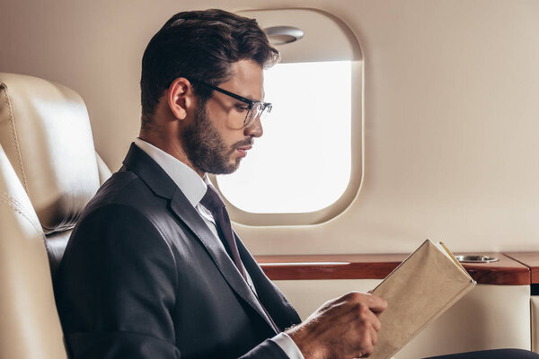 вид сбоку на красивого бизнесмена в костюме, читающего книгу в частном самолете
 