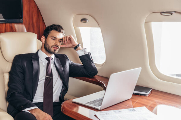красивый бизнесмен в костюме смотрит на ноутбук в частном самолете
 