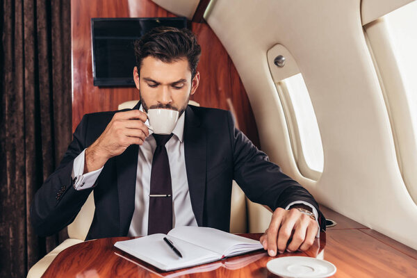 красивый бизнесмен в костюме пьет кофе в частном самолете
 