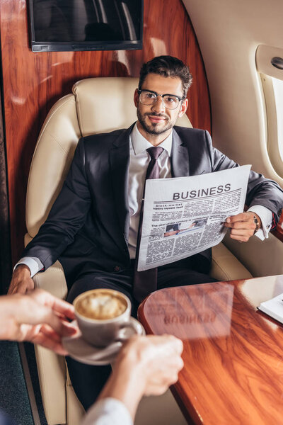 cropped view of flight attendant giving cup of coffee to handsome businessman in suit in private plane 