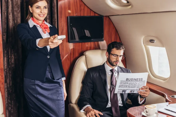 英俊的商人穿着西服 一边看报纸 一边笑着在私人飞机上牵着遥控器的空姐 — 图库照片