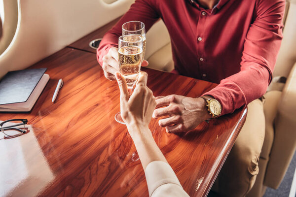 cropped view of boyfriend and girlfriend clinking with champagne glasses in private plane 