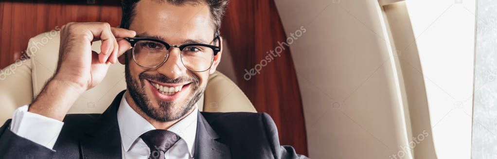 panoramic shot of handsome businessman in glasses smiling and looking at camera in private plane 