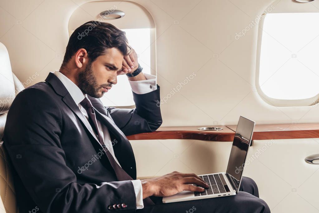 side view of handsome businessman in suit using laptop in private plane 