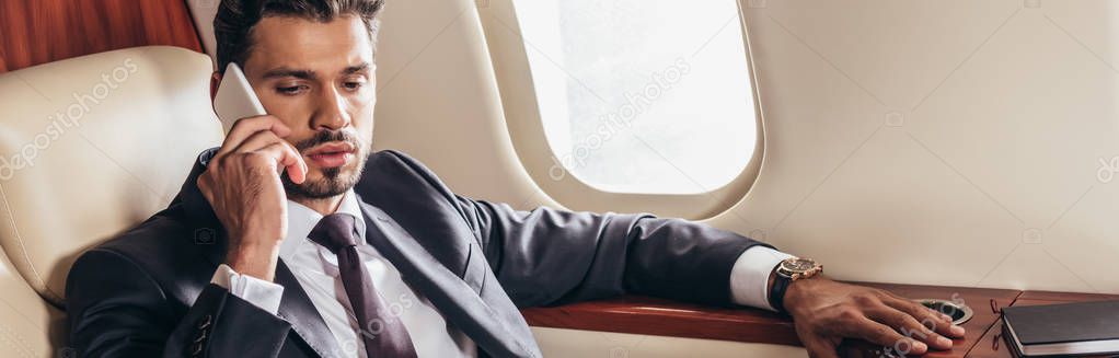 panoramic shot of handsome businessman in suit talking on smartphone in private plane 