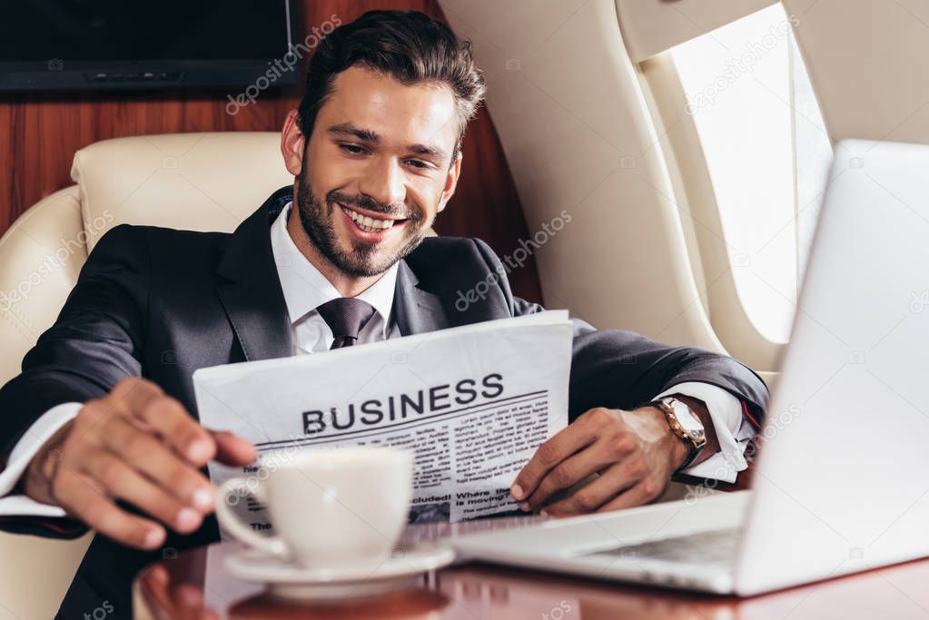 selective focus of smiling businessman in suit with newspaper taking cup in private plane 