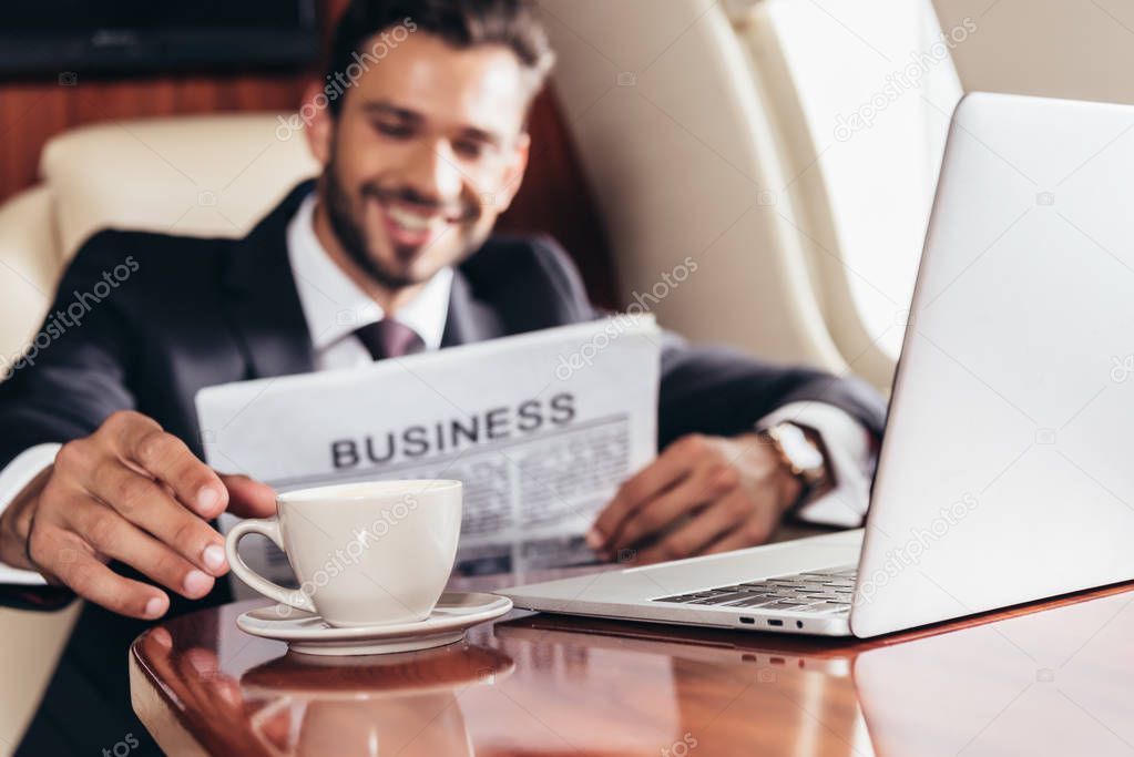 selective focus of smiling businessman in suit with newspaper taking cup in private plane 