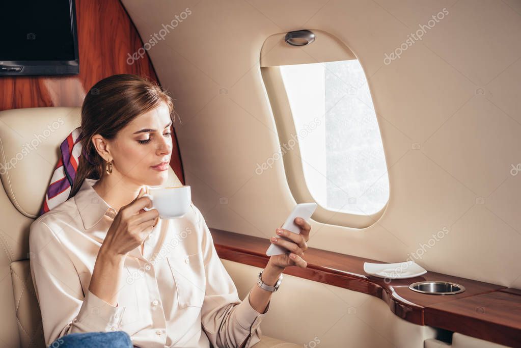 attractive woman in shirt holding cup and using smartphone in private plane 