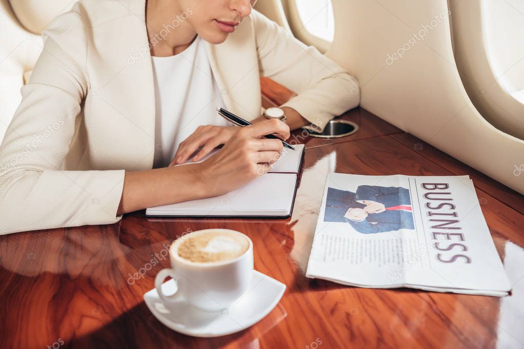 cropped view of businesswoman in suit writing in notebook in private plane 