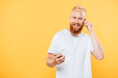 Gülümseyen sakallı adam kulaklıklı ve akıllı telefonlu müzik dinliyor, sarıda izole edilmiş.