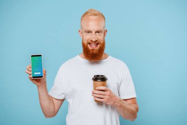 Kyiv, Ukrayna - 27 Ağustos 2019: sakallı ve kahveli bir adam twitter uygulamalı akıllı telefonu göstermek için mavi üzerine izole edilmiş.